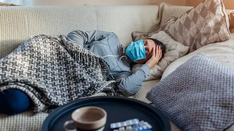 Sick woman having flu or cold. Girl lying in bed having headache wearing protective mask by pills and water on table. (Sick woman having flu or cold. Girl lying in bed having headache wearing protective mask by pills and water on table., ASCII, 118 co